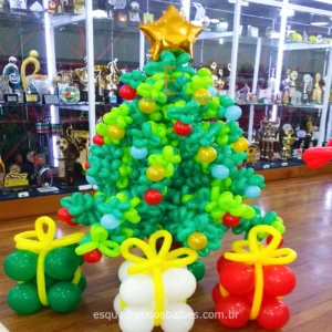 A imagem trata-se de uma arvore de natal confeccionada em balões em tons de verde, complementando a decoração três caixas de presentes feitas de balões nas cores verde, branco e vermelho.