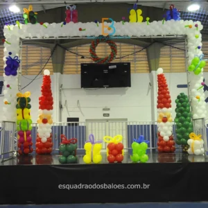 decoração de balões para festa natalina, a imagem apresenta esculturas de balões que simulam arvores de natal, caixas de presentes, Papai Noel, guirlanda natalina e uma moldura de balões brancos compondo o cenário.